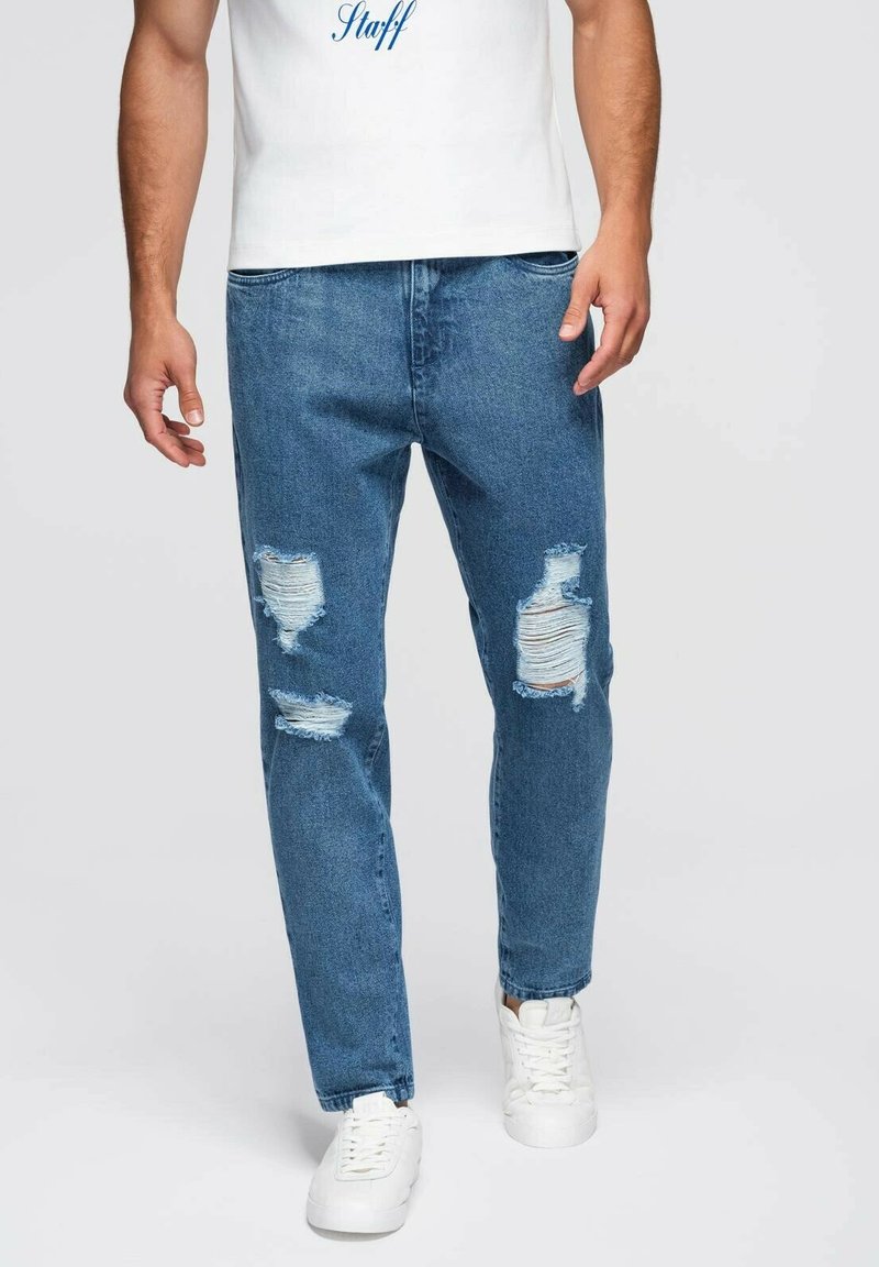 Jeans de mezclilla azul desgastados con detalles rasgados y corte recto, combinados con zapatillas blancas y una camiseta blanca con texto azul.
