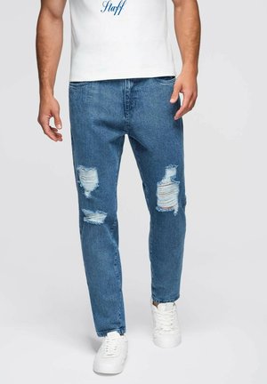 Abgenutzte blaue Jeans mit Rissdetails und geradem Schnitt, kombiniert mit weißen Sneakers und einem weißen Hemd mit blauer Schrift.