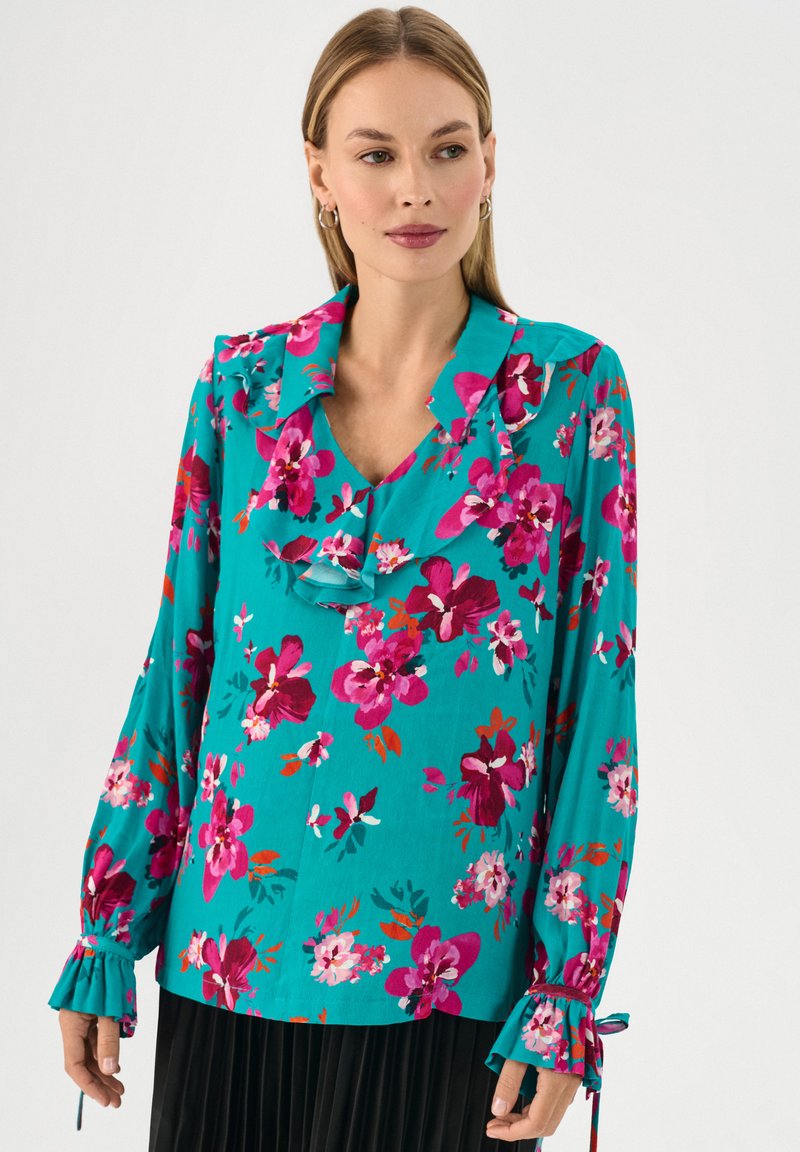 Blusa floreale in tessuto turchese, con colletto voluttuoso, maniche lunghe con dettagli a fiocco e un vivace mix di fiori rosa e bianchi.