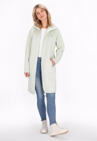 Cappotto lungo verde chiaro con zip e cappuccio; texture morbida, tasche laterali e zip bianca a contrasto. Abbinato a jeans blu e scarpe chiare.