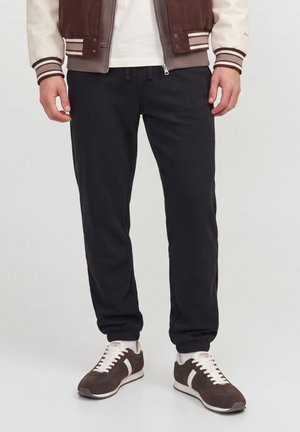 Mand iført sorte joggingbukser med elastiske manchetter, brune og hvide sneakers, en hvid skjorte og en brun og cremefarvet varsityjakke.