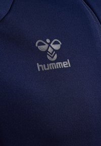 Marineblauwe stof met een zilveren Hummel-logo, met een gestileerde hommel boven de merknaam op sportkledingmateriaal.