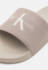 Sive tekstilne papuče s širokom trakom koja ima bijeli "CK" logo i tekst "Calvin Klein Jeans", s potplatom koji nalikuje gumi.