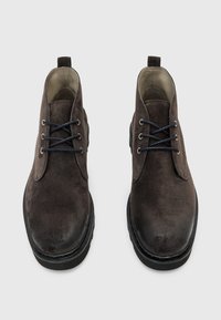 Stivali ankle in suede marrone con texture liscia, punta rotonda e lacci scuri a contrasto. Presentano una robusta suola in gomma nera.