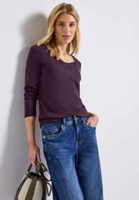 Mörklila långärmad topp med fyrkantig halsringning, tillsammans med ljusblå denimjeans. En grå handväska med gröna accenter hålls i ena handen.