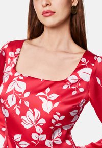 Rotes Satin-Kleid mit quadratischem Ausschnitt und langen Ärmeln, verziert mit weißen Blumenmustern. Glatte Textur mit subtiler Seideneffekt.