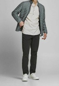 Muž ve šedé proševané bundě přes béžové tričko typu henley, černých kalhotách a bílých teniskách, stojící na jednobarevném šedém pozadí.