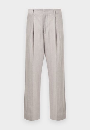 Junger Mann steht mit der Hand in der Tasche, trägt eine hellgraue Reißverschlussjacke, ein schwarzes Hemd, beige weite Hosen und schwarze Schuhe.