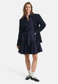 Rochie din bumbac bleu marin cu fustă în straturi și talie evidențiată cu un brâu, stratificată sub un sacou texturat de culoare închisă. Pantofi negri cu talpă joasă, cu accente decorative.