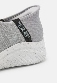 Baskets slip-on grises avec une tige texturée et une étiquette noire, présente une semelle blanche tachetée et un design arrondi.