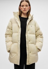 Beige puffjacka med kviltat mönster, hög krage, framdragkedja och tryckknappar. Har en liten logotyp på bröstet och sidofickor.