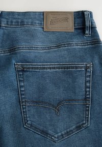 Jeans de mezclilla azul oscuro con un bolsillo trasero cosido, etiqueta de cuero con la marca y detalles de textura sutiles.