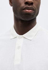 Κοντινό πλάνο ενός άνδρα με σκούρο χρώμα δέρματος που φοράει λευκό πόλο με γιακά και δύο κουμπιά κλειστά, με φόντο μονόχρωμο.