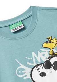 Hellblaues T-Shirt mit Snoopy, der eine Sonnenbrille trägt, und Woodstock, der auf seinem Kopf steht. Das Peanuts-Label ist im Kragen sichtbar.