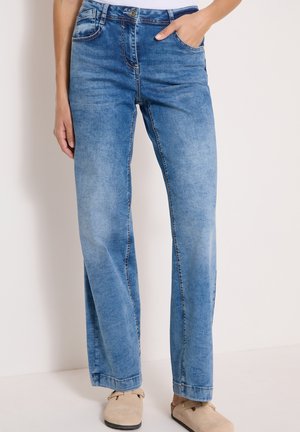 Straight-leg blå jeans med en falmet vask, båret med beige slip-on sko, en hånd i lommen, hvid baggrund.