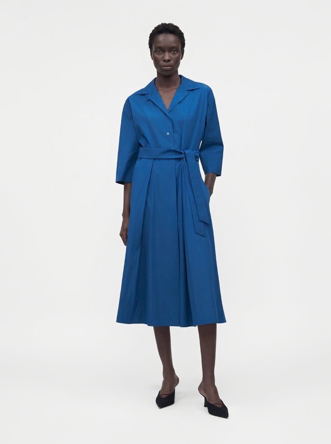 Femme portant une robe bleue longue jusqu'aux genoux, manches longues, avec col, boutonnière devant, ceinture à la taille et talons noirs à bouts pointus, debout devant un fond uni.