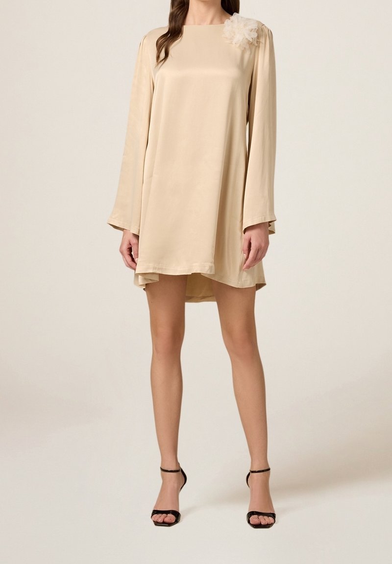 Žena v béžových mini šatech s dlouhým rukávem a květinovou ozdobou na rameni, se černými lodičkami na kotníku.