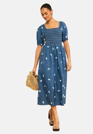 Frau trägt ein blaues, geblümtes Midikleid mit Puffärmeln und gerafftem Oberteil, hält eine geflochtene Handtasche und trägt offene Plateau-Sandaletten.