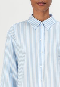 Camicia button-up a righe blu chiaro e bianche con collo classico, realizzata in tessuto morbido, dotata di pattina frontale e maniche lunghe.