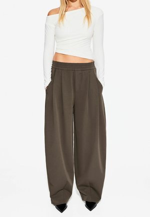 Kobieta ubrana w biały, asymetryczny, odkrywający ramię, długi crop top z rękawami oraz luźne, szerokie, ciemnobrązowe spodnie z czarnymi, spiczastymi butami.