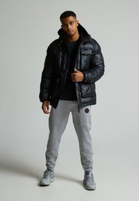 Giacca a vento nera con finish lucido, cappuccio e tasche frontali; abbinata a pantaloni della tuta grigi con toppe sulle ginocchia e un logo.