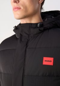 Giacca a vento nera con collo alto e cappuccio, con un'etichetta rettangolare rossa "HUGO". Tessuto liscio con sezioni trapuntate e toggle regolabili.