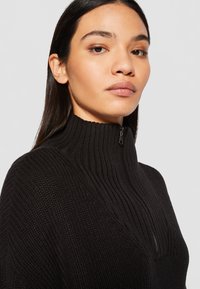 Zwarte gebreide sweater met een hoge kraag, met een korte rits en een gestructureerd patroon. Gladde afwerking rond de halslijn en schouders.