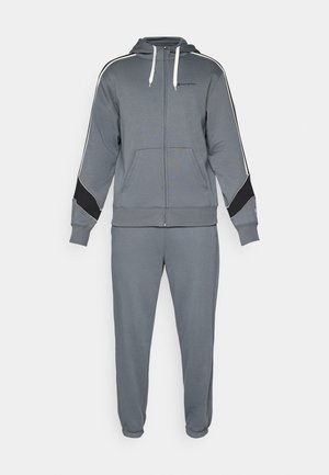 Tuta grigio con zip e cappuccio, caratterizzato da dettagli bianchi e neri. Il tessuto è liscio e presenta due tasche frontali sulla giacca e polsini elasticizzati.