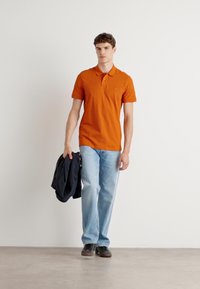 Oransje polo med en liten lommene logo, kombinert med lyseblå jeans. Modellen holder en mørk jakke og står på en nøytral bakgrunn.