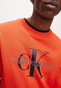 Pomarańczowa bluza z kapturem z czarnym, haftowanym logo "CK" oraz napisem "CALVIN KLEIN JEANS". Czarny ściągacz przy dekolcie; widoczna faktura bawełny.