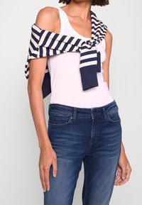 Mujer vistiendo una camiseta sin mangas blanca y jeans azul oscuro, con un suéter a rayas azul marino y blanco sobre los hombros y atado en el pecho.