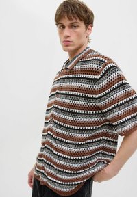 Chemise à manches courtes avec col, présentant un motif rayé multicolore en marron, noir et blanc, fabriquée en tissu léger texturé avec des boutons.