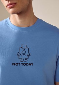 Ljusblå kortärmad t-shirt med en svart tryckt tecknad karaktär med höghatt som ser sur ut, och texten "IKVÄL" nedanför.