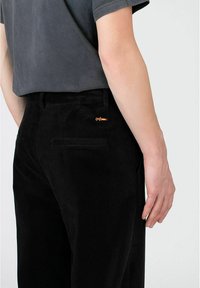 Svart kordfløjelsbukser med lodrette ribber, der har en baglomme og en orange logo-detalje på højre side nær taljen.