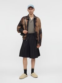 Homme portant une casquette bleue, une veste marron à motifs, une chemise grise, une jupe plissée noire, des chaussettes noires, des chaussons beiges, et portant un sac à bandoulière noir.