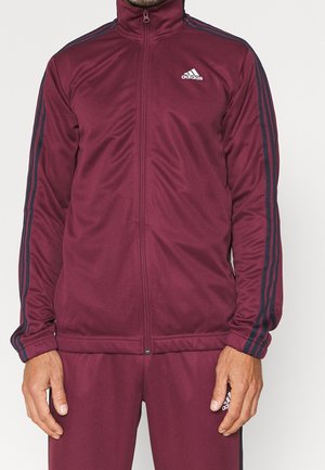 Person trägt einen kastanienbraunen Adidas-Trainingsanzug mit schwarzen Streifen auf Ärmeln und Hosen, stehend vor einem schlichten hellen Hintergrund.