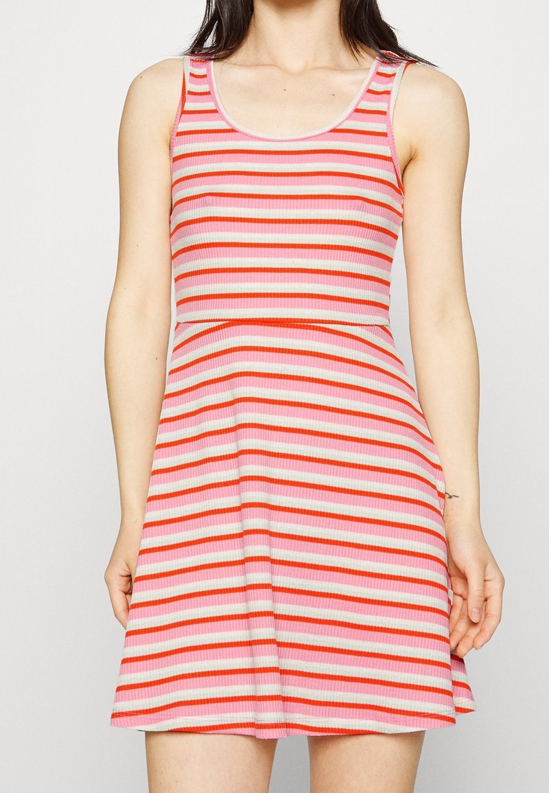 Femme portant une robe sans manches à encolure dégagée avec des rayures horizontales roses, rouges et blanches, debout devant un fond uni.