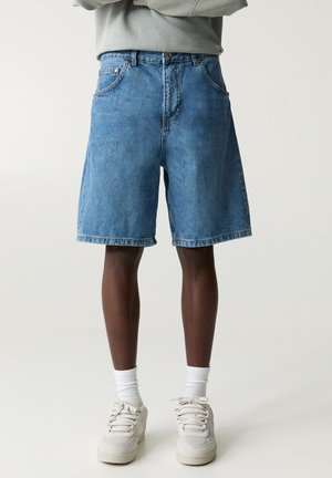 Persona che indossa pantaloncini di jeans blu, calzini bianchi e scarpe da ginnastica bianche, in piedi con le braccia incrociate davanti a uno sfondo chiaro e semplice.