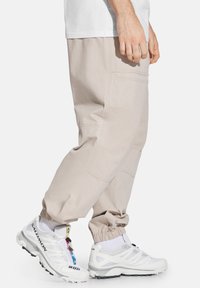 Beżowe, zwężane spodnie cargo z elastycznymi mankietami, posiadające wiele kieszeni i przeszycia. Noszone z białymi butami sportowymi i skarpetkami.