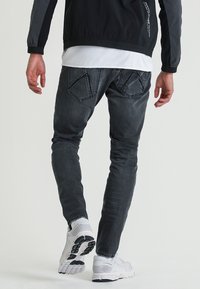 Jean gris foncé coupe slim, avec des poches arrière distinctes en forme de losange, associé à des chaussures de sport blanches et une veste noire.