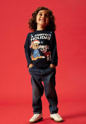 Langärmliges, marineblaues Hemd mit Cartoon-Hunden, die Feiertagsaccessoires tragen, kombiniert mit blauen Jeans und weißen Sneakers. Roter Hintergrund.