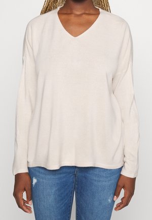 Personne à la peau foncée avec des cheveux tressés portant un pull crème à col en V et manches longues, ainsi qu'un jean bleu déchiré.