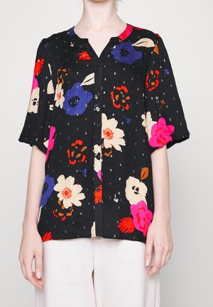 Blouse noire à boutons avec de grandes impressions florales colorées en rose, rouge, violet et beige, portée avec un pantalon clair, visible du cou aux hanches.