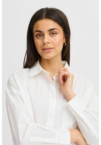 Camicia bianca con colletto classico, texture morbida e maniche lunghe. Il tessuto appare leggero e leggermente trasparente.