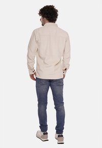 Camicia di velluto a coste beige con yoke posteriore e polsini abbottonati, abbinata a jeans di denim blu e sneakers di colore chiaro.