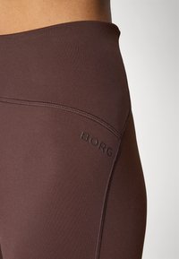 Braune sportliche Leggings verfügen über ein nahtloses Design, eine Logodetail auf dem Bund und eine glatte Textur. Die sichtbaren Nähte setzen Akzente.