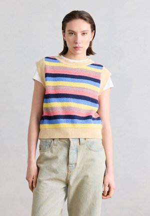 Donna che indossa un gilet senza maniche in maglia con strisce orizzontali gialle, rosa, blu e blu scuro sopra una T-shirt bianca e jeans beige chiaro.