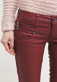 Burgundy slim jeans med en slät yta, med en dragkedjeficka och dekorativ sömnad. Metallaccenter är synliga.