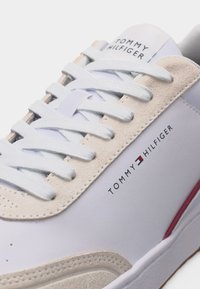 Λευκό και μπεζ αθλητικό παπούτσι Tommy Hilfiger με λευκά κορδόνια, λογότυπο της μάρκας στο πλάι και στη γλώσσα, και κόκκινο τόνισμα στη σόλα.