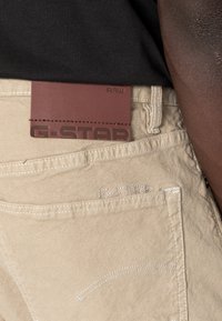 Calças de algodão bege com costura branca, apresentando uma etiqueta de marca em couro castanho que diz "G-STAR RAW" na cintura.